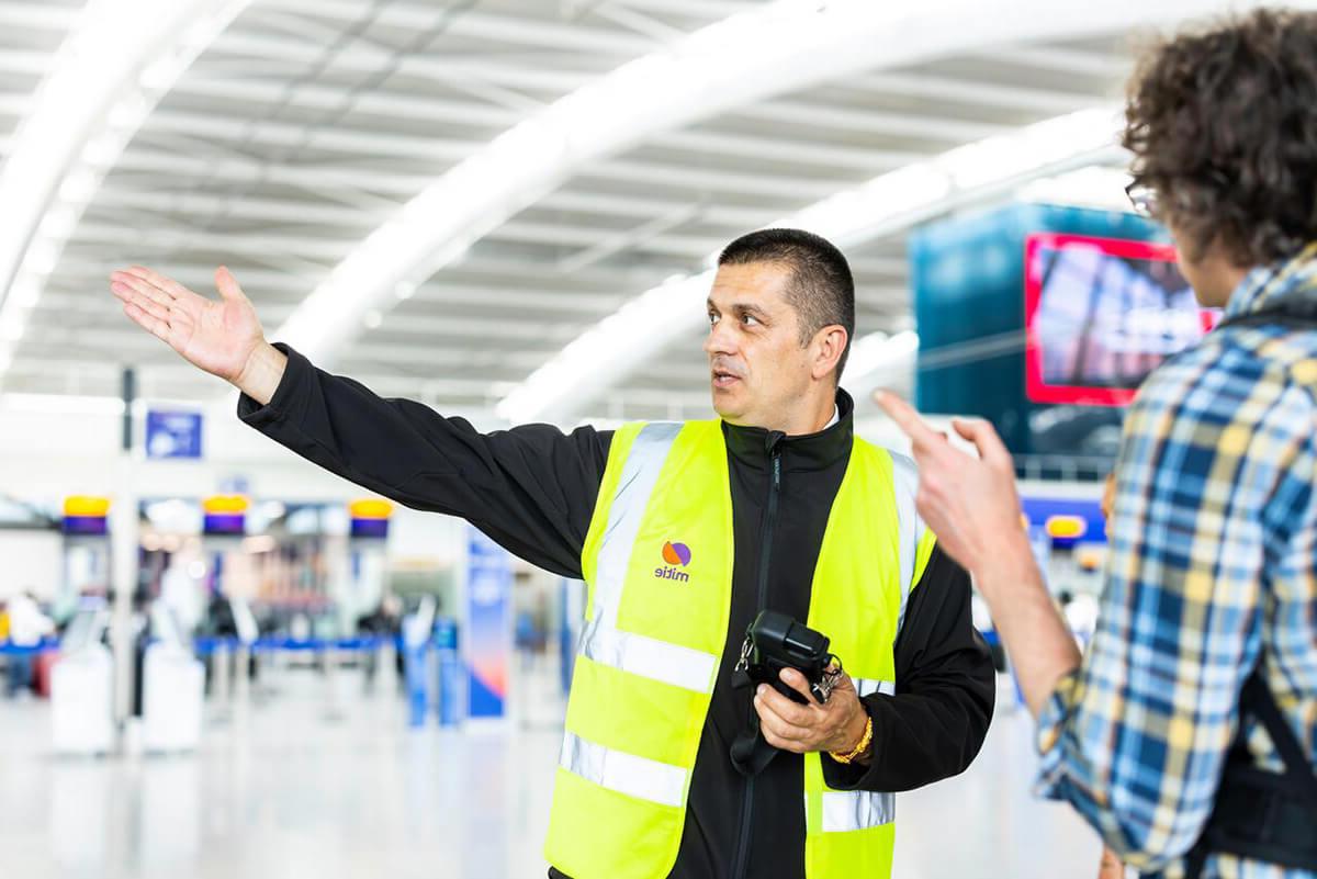 Male security guard at Heathrow Airport, wearing a gpk电子 branded high vis, pointing to the right and directing a customer